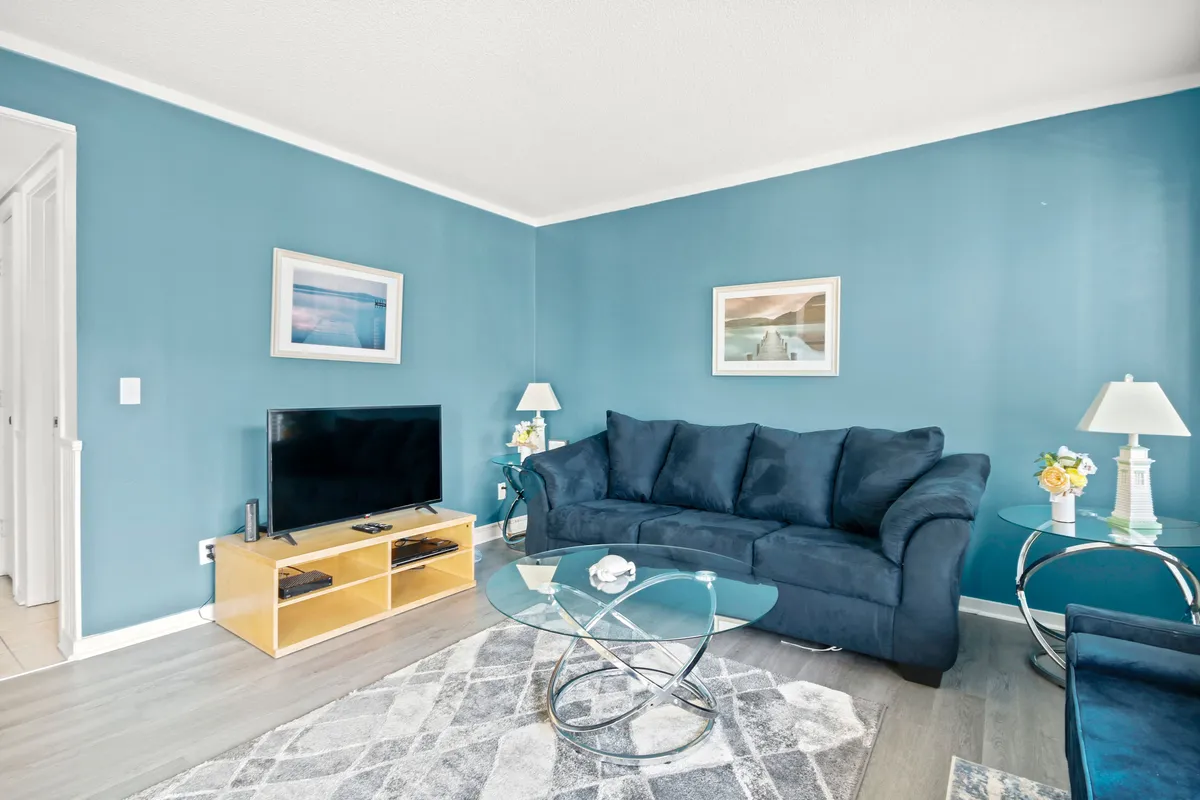 A welcoming living room with a plush blue sofa, glass coffee table, television, decorative lamps, and wall art. The seating arrangement is designed for relaxation and conversation, offering a cozy spot for gathering after a day at the beach.