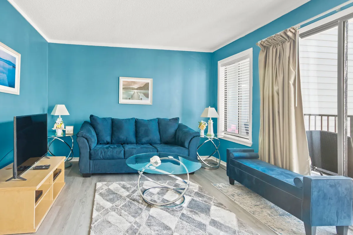 The image shows a bright, inviting living room featuring a plush blue couch, a modern glass coffee table, a decorative painting above the couch, and a sleek TV stand. The calm, coastal-inspired decor is perfect for relaxation or socializing, with natural light enhancing the welcoming atmosphere.
