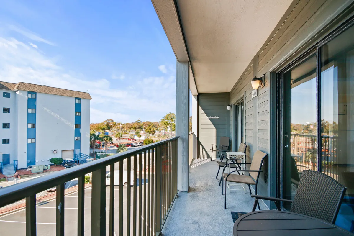 A private balcony on the third floor with comfortable seating overlooks the nearby area, with hints of the ocean visible. Morning light spills onto the balcony, suggesting a peaceful setting ideal for enjoying coffee or an evening drink. The scene exudes a casual, laid-back mood that aligns with a romantic or relaxing getaway.