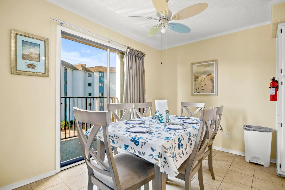 A dining area with a floral tablecloth on the dining table, a ceiling fan, a window inviting sunlight, and tasteful decorative art. The space feels intimate yet open, ideal for shared meals or planning vacation adventures.