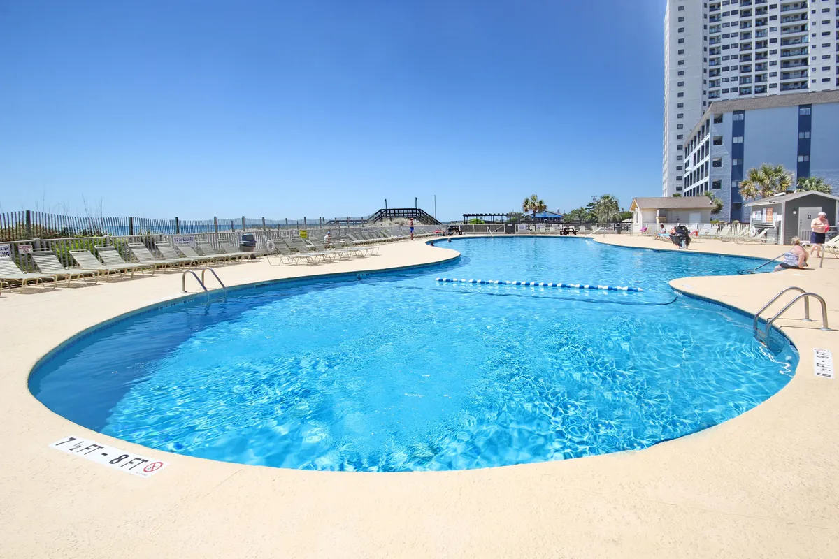 A lively resort pool scene featuring shimmering blue water, lounge chairs arranged around the deck, and water features. The area is surrounded by resort buildings, and the setting sun casts a warm glow, hinting at the social and recreational opportunities for groups and families.