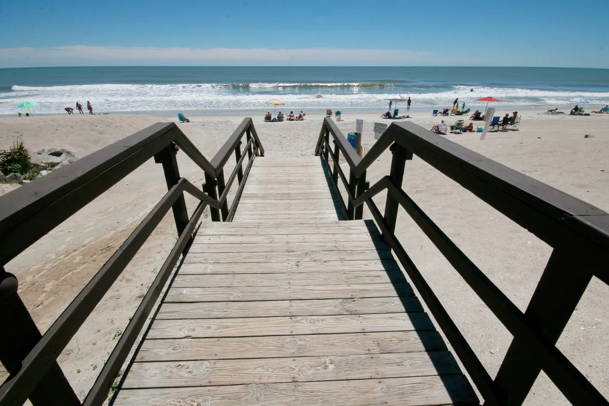 A wide, sun-drenched beach lined with beach umbrellas and sunbathers. A wooden walkway leads directly onto the sandy shoreline, with gentle waves rolling in and people engaging in various beach activities such as swimming, picnicking, and playing beach games.