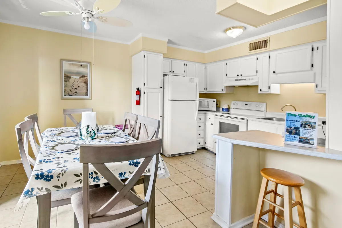The kitchen and adjacent dining table are visible, with a floral tablecloth, kitchen appliances in the background, and warm wall art. The scene suggests an inviting space for group meals and shared celebrations.