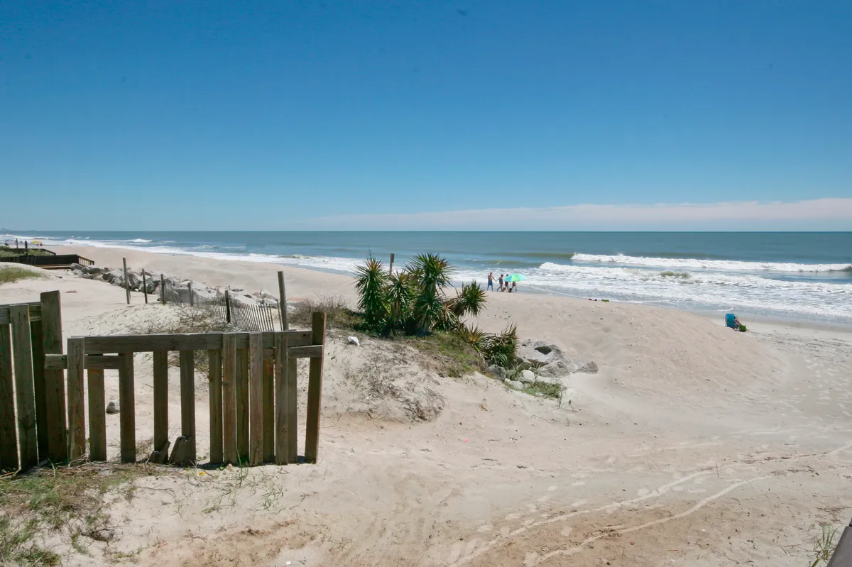 A serene view of the beach and ocean, framed by palm plants and a wooden fence, with people strolling, swimming, and sunbathing. The shoreline and gentle waves highlight the resort's direct beach access.