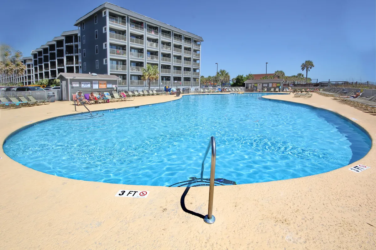 A sparkling outdoor pool surrounded by lounge chairs and palm trees, with a residential building in the background. The pool area is bathed in soft light, inviting visitors to relax, swim, or socialize. The relaxed atmosphere is perfect for unwinding or enjoying a sunset swim after a day on the beach.