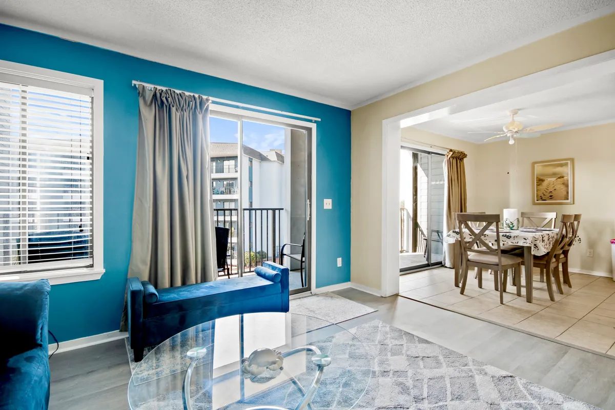 An airy, open-concept space featuring a blue accent wall, a glass coffee table, a dining table, and large curtained windows that fill the room with natural light. The design encourages interaction and shared moments, with seamless flow between living and dining areas.