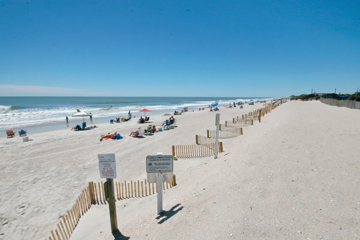 A wide, sandy stretch of Myrtle Beach lined with umbrellas and people lounging near the gentle waves. The scene includes a wooden fence and a shoreline perfect for walking, sunbathing, or watching the sunset, evoking a leisurely and carefree mood ideal for romantic strolls or relaxing with friends.