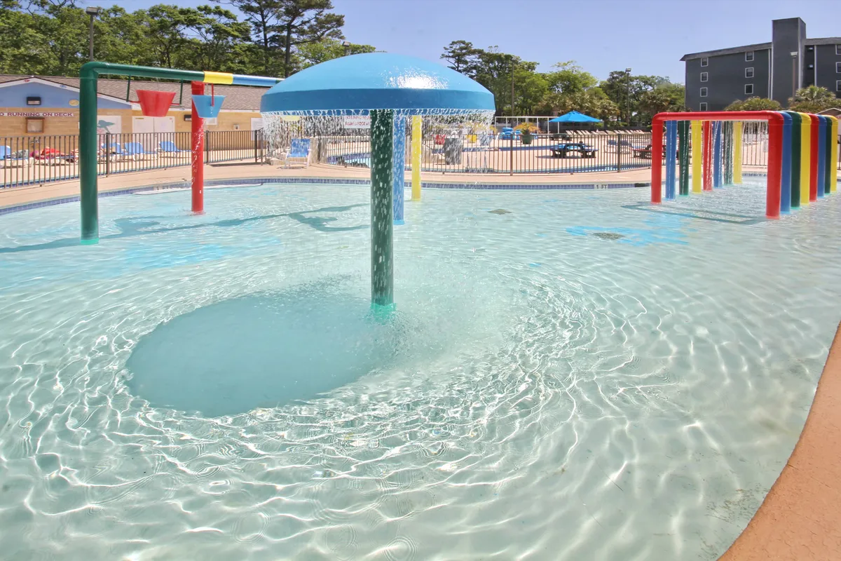 A vibrant scene featuring a splash pad with colorful water features and clear blue water. Children are joyfully playing under streams and sprays, while families are gathered around enjoying the sunshine and relaxed atmosphere. The playful design elements and cool water make the area inviting for kids of all ages.
