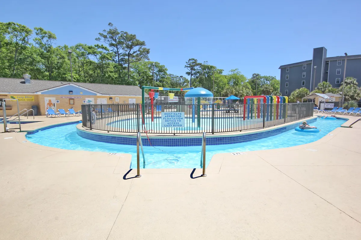 A lively outdoor scene showing a sparkling swimming pool with colorful water slides and plenty of lounge chairs nearby. Children are seen playing and sliding, while families relax poolside, soaking up the sun on a beautiful day. The bright ambiance radiates joy and excitement.